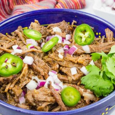 Shredded beef topped with sliced jalapeรฑos, chopped red onion, and cilantro in a blue bowlโperfect for hearty recipesโset against a colorful striped cloth and diced onions in the background.