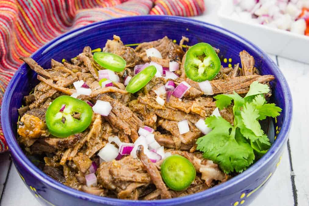 Shredded beef topped with sliced jalapeños, chopped red onion, and cilantro in a blue bowl—perfect for hearty recipes—set against a colorful striped cloth and diced onions in the background.