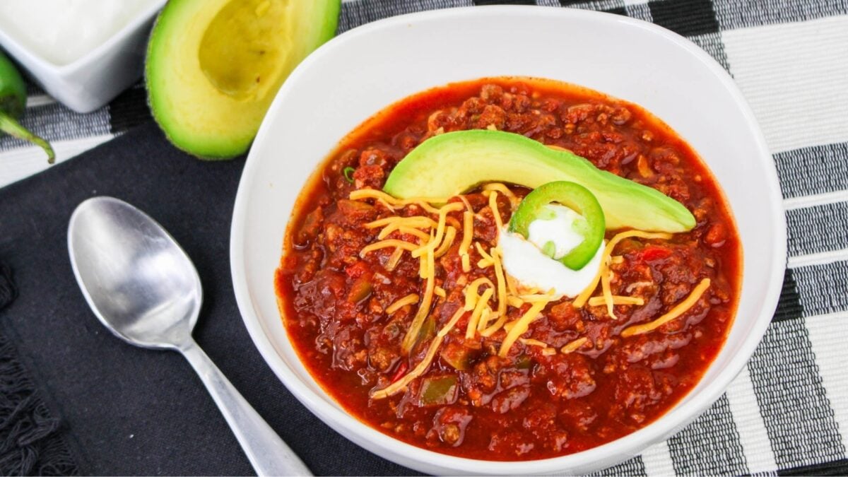 A bowl of chili topped with shredded cheese, avocado slices, a dollop of sour cream, and a jalapeño slice—served with a spoon and halved avocado—is perfect for hearty recipes and cozy winter meals.