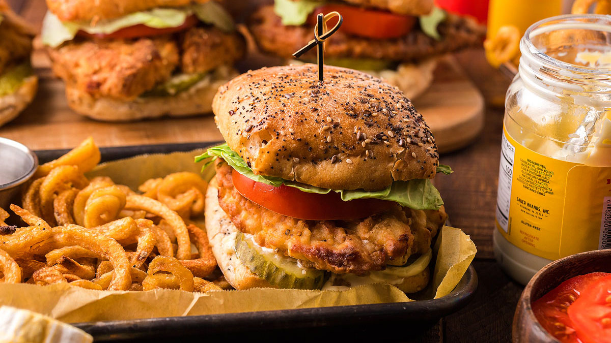 A breaded chicken sandwich with crispy texture, topped with lettuce, tomato, and pickles on a seeded bun served with curly fries on a tray; a jar of mustard is nearby—perfect inspiration for your own fried chicken recipes.