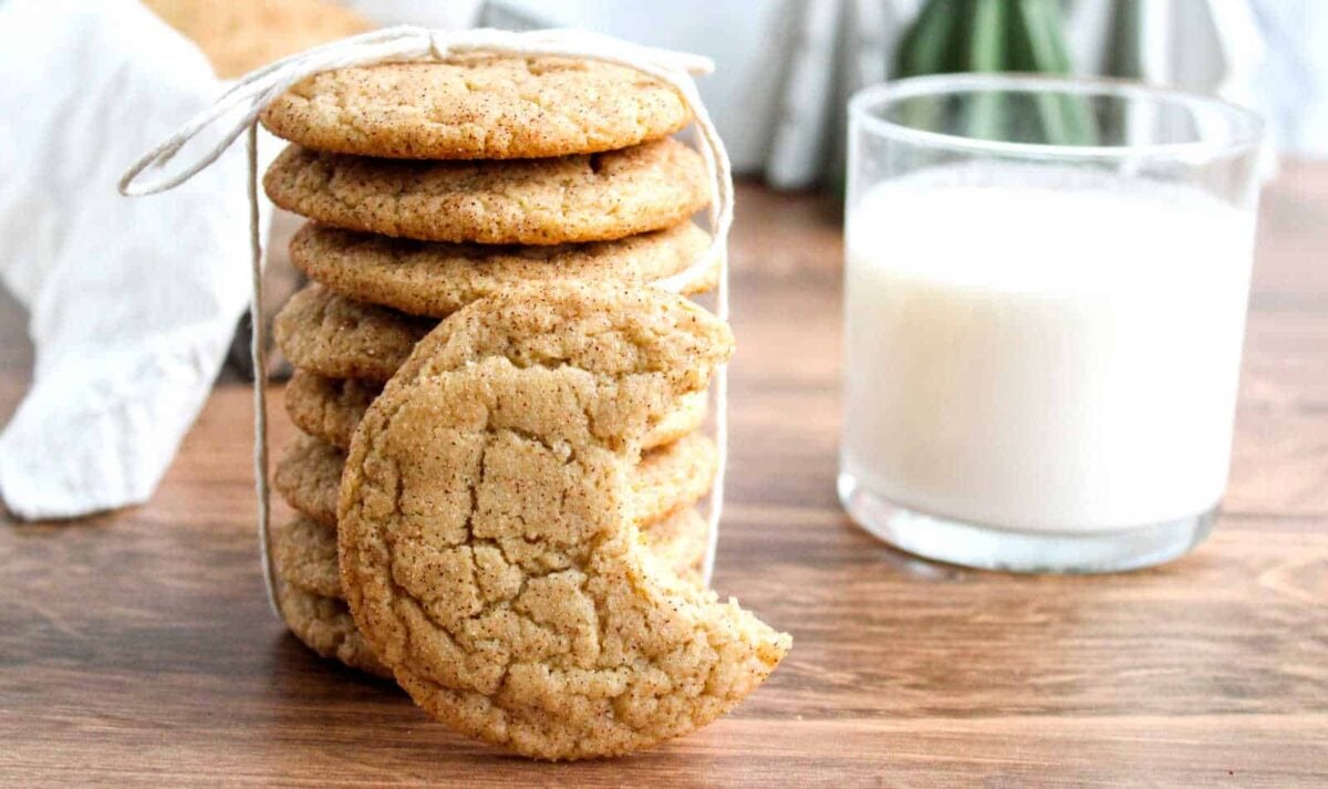 A stack of firm snickerdoodle cookies tied with string sits on a wooden surface next to a glass of milk; one sweet cookie has a bite taken out. Perfect for those seeking classic snickerdoodle recipes.
