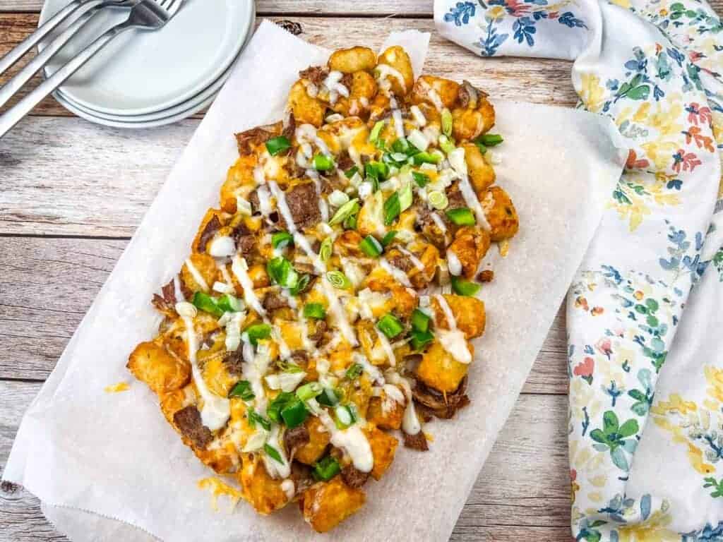 A tray of tater tots topped with melted cheese, chopped green onions, and sauce served on parchment paper delivers crisp flavor&mdash;perfect as Air Fryer Appetizers without the oil, alongside plates, forks, and a floral napkin.