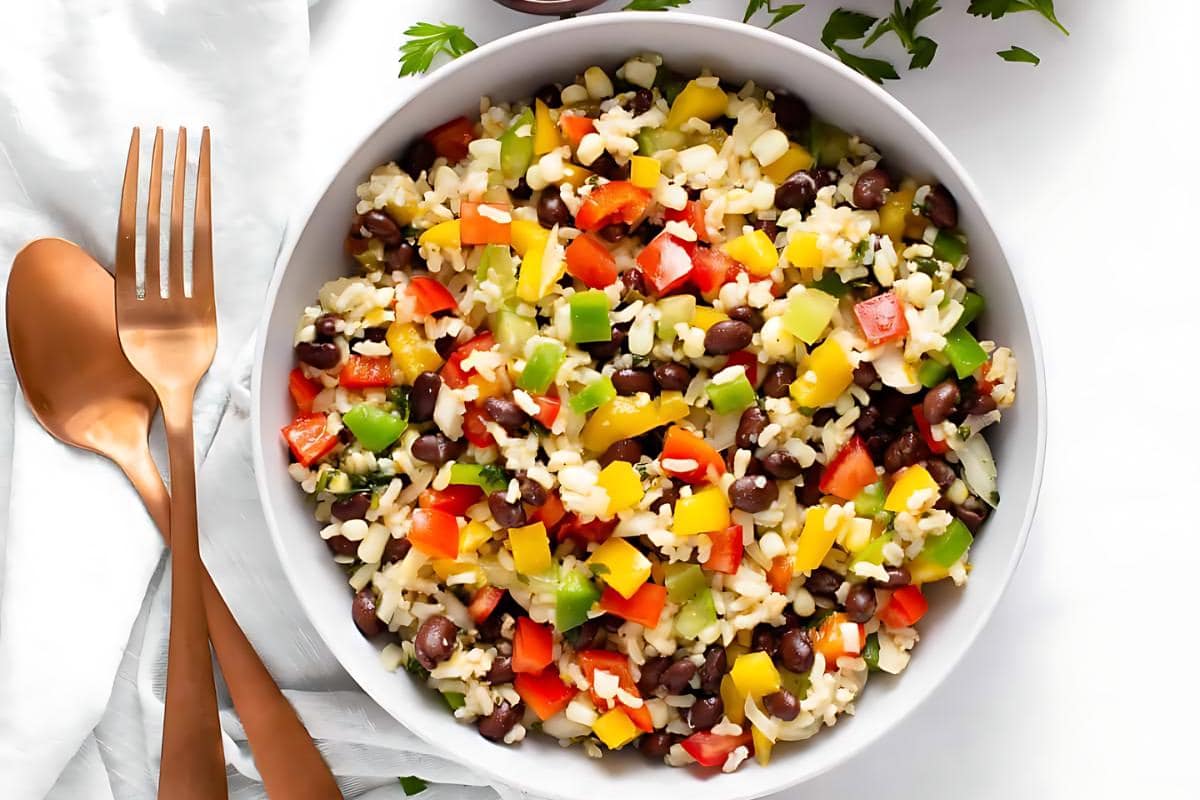 A bowl of rice salad with black beans, chopped red, yellow, and green bell peppers, and diced cucumber makes a colorful addition to your side dishes collection, placed next to two bronze-colored utensils on a white surface.