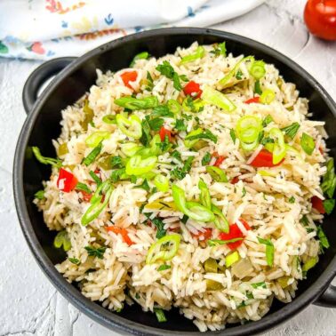 A black bowl filled with cooked rice mixed with chopped green onions, red bell peppers, and herbs sits on a white surface—perfect for adding to your collection of flavorful rice side dishes or recipes.