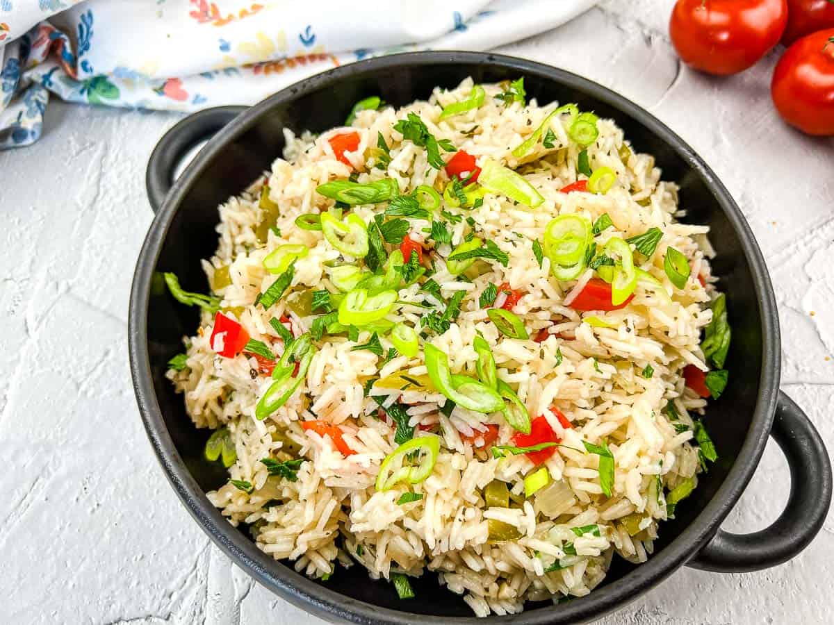 A black bowl filled with cooked rice mixed with chopped green onions, red bell peppers, and herbs sits on a white surface&mdash;perfect for adding to your collection of flavorful rice side dishes or recipes.