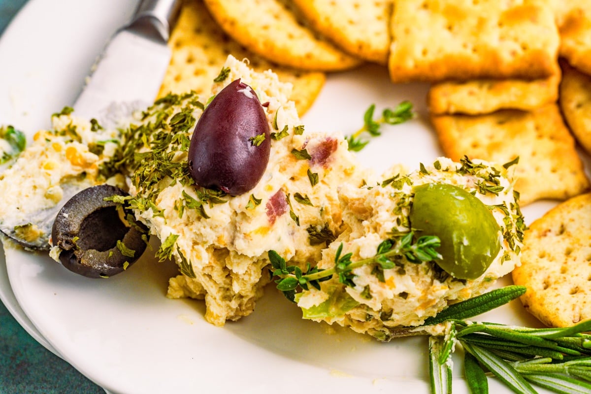A plate with herbed cheese spread garnished with black and green olives, fresh herbs, crackers, and a butter knife.