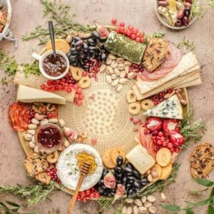 A round wooden board with assorted cheeses, crackers, meats, grapes, figs, nuts, and jams arranged decoratively, with herbs and a small bowl of olives nearby.
