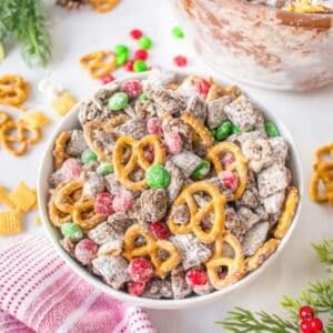 A bowl of holiday snack mix with pretzels, chocolate-covered cereal, red and green candy-coated chocolates, and powdered sugar on a white surface.