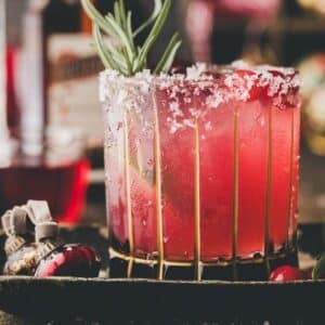 A red cocktail in a glass with a salted rim, garnished with a sprig of rosemary and lime, sits on a tray with cranberries and a festive ornament nearby.