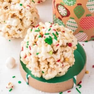 A popcorn ball decorated with colorful holiday sprinkles sits on a green-lined round cardboard lid, with a festive container and more popcorn balls in the background.