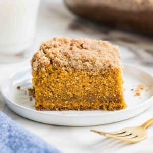 A slice of pumpkin coffee cake topped with cinnamon crumb streusel on a white plate, with a gold fork and a glass of milk nearby.