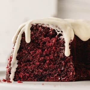 A close-up of a slice of red velvet cake with white icing dripping over the top, set on a white surface.