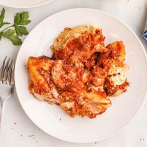 A white plate with stuffed pasta shells topped with tomato sauce and grated cheese, garnished with herbs. A fork and parsley are on the side.