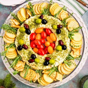 A round platter with a cheese ball arranged in a wreath shape, topped with olives and cherry tomatoes, surrounded by crackers and sprigs of fresh herbs.