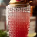 A hand with pink nail polish holds the lid of a textured glass cocktail shaker filled with a red and pink drink.