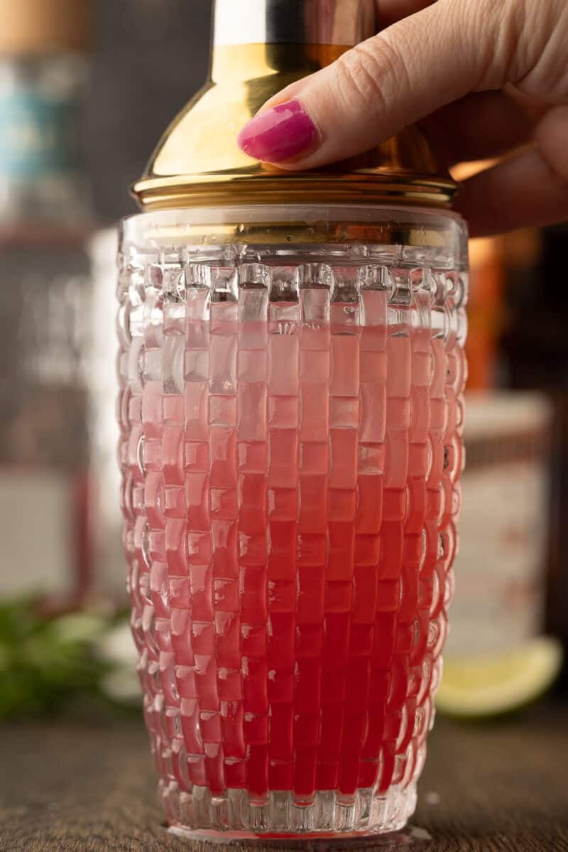 A hand with pink nail polish holds the lid of a textured glass cocktail shaker filled with a red and pink drink.