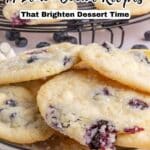 A close-up of lemon cookies with blueberries on a plate and cooling rack, topped with coarse sugar. Text reads "14 Lemon Cookie Recipes That Brighten Dessert Time.