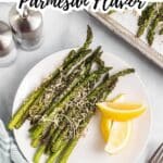 A plate of roasted asparagus topped with grated parmesan cheese, served with lemon wedges, next to a tray of asparagus and salt and pepper shakers.