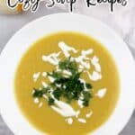 A bowl of creamy yellow soup topped with white drizzle and chopped herbs, placed on a white plate with part of an apple and bread visible in the background. Text reads "15 Bowls of Warmth Powered by Cozy Soup Recipes.