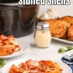 A plate of stuffed pasta shells with tomato sauce and cheese sits on a table, with garlic bread, a cheese shaker, and a slow cooker in the background.