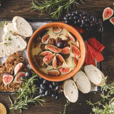 A dish of baked brie topped with figs and pecans is surrounded by sliced bread, crackers, grapes, fresh figs, and herbs on a wooden table.