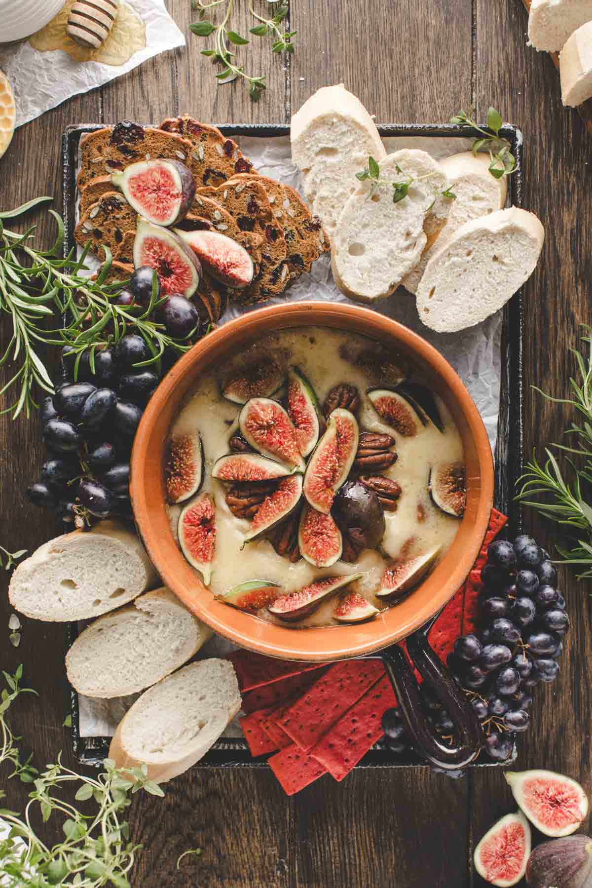 A baked brie topped with fresh figs and pecans surrounded by sliced bread, grapes, and herbs on a wooden table.