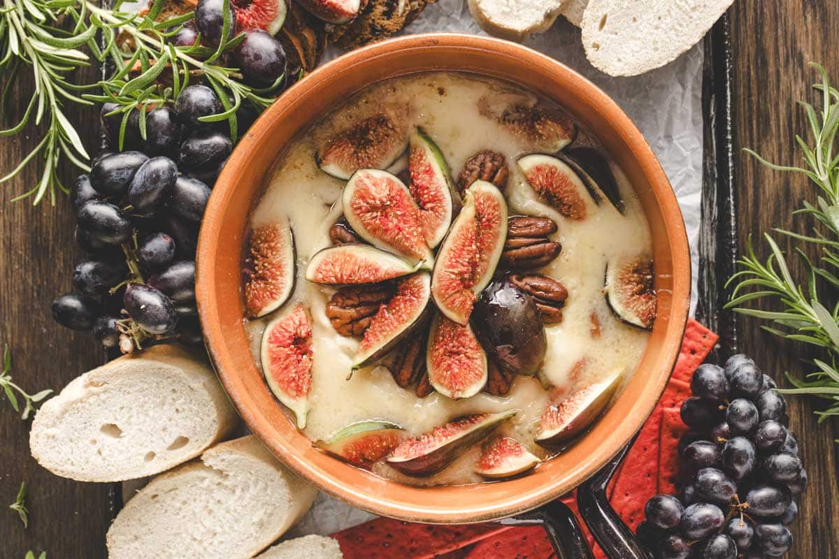 A baked brie topped with sliced figs and pecans in a dish, surrounded by clusters of grapes, rosemary sprigs, and slices of white bread on a wooden table.
