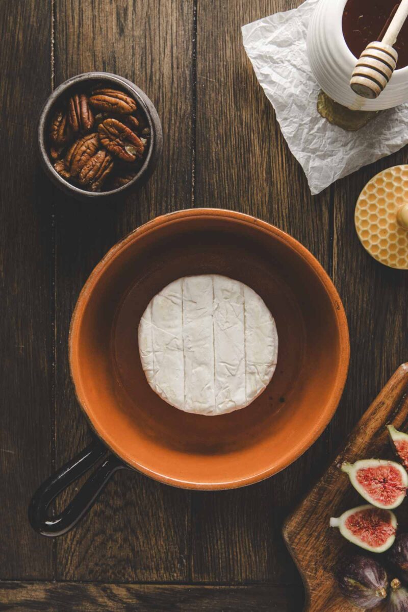 A round wheel of brie cheese sits in a brown baking dish next to a bowl of pecans, honey with a dipper, and a cutting board with sliced figs on a wooden table.