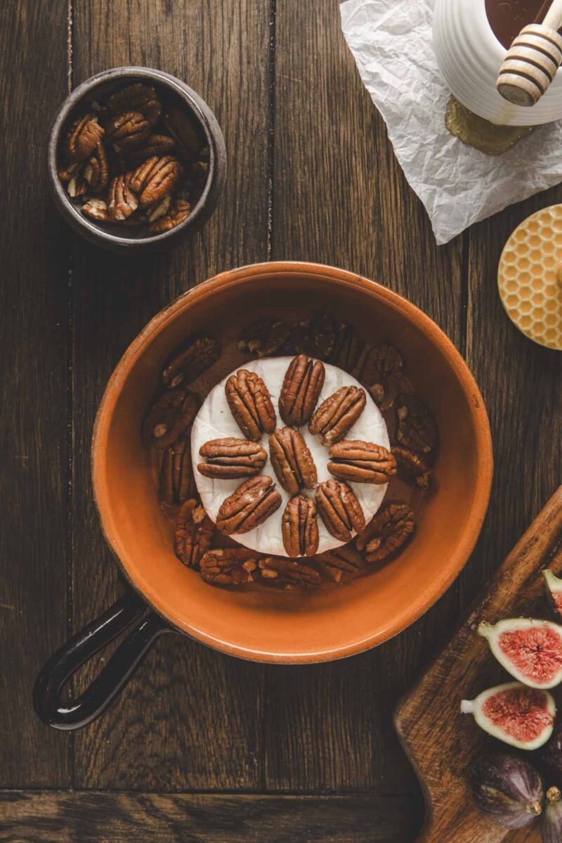 A round of brie cheese topped with pecans in a brown dish, next to a bowl of pecans, honey, and a wooden board with figs.