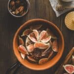A ceramic pan filled with sliced figs and pecans on a wooden table, with a bowl of pecans, honey dipper, and a mug nearby.
