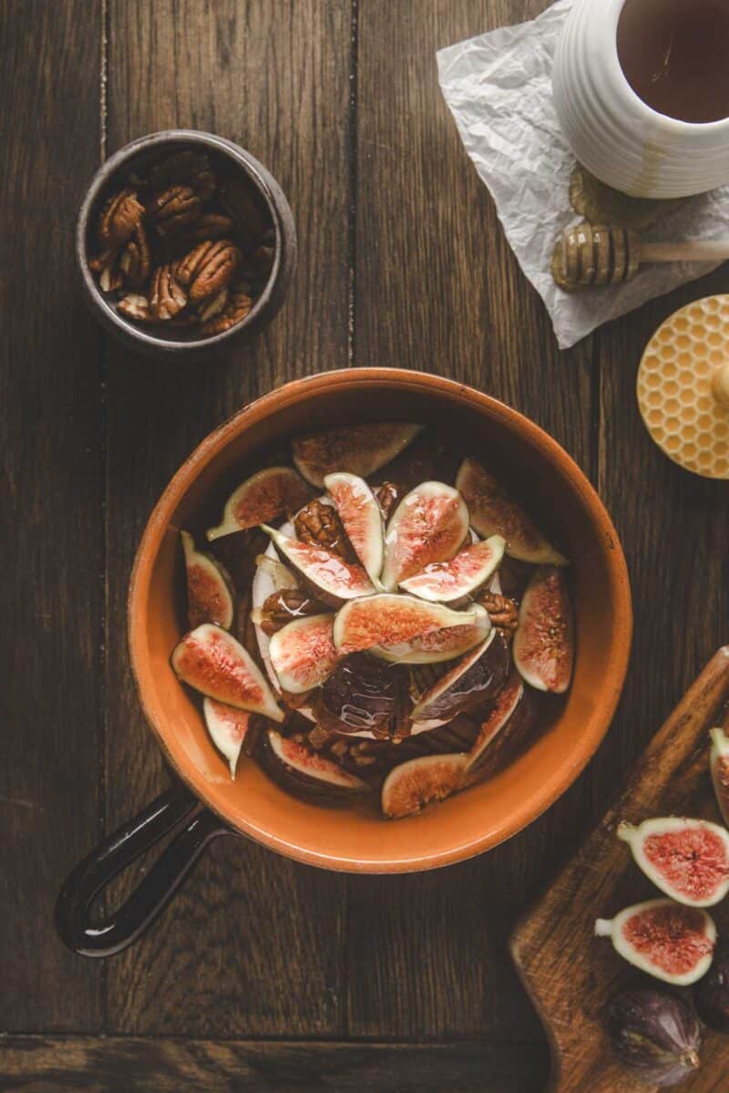 A ceramic pan filled with sliced figs and pecans on a wooden table, with a bowl of pecans, honey dipper, and a mug nearby.