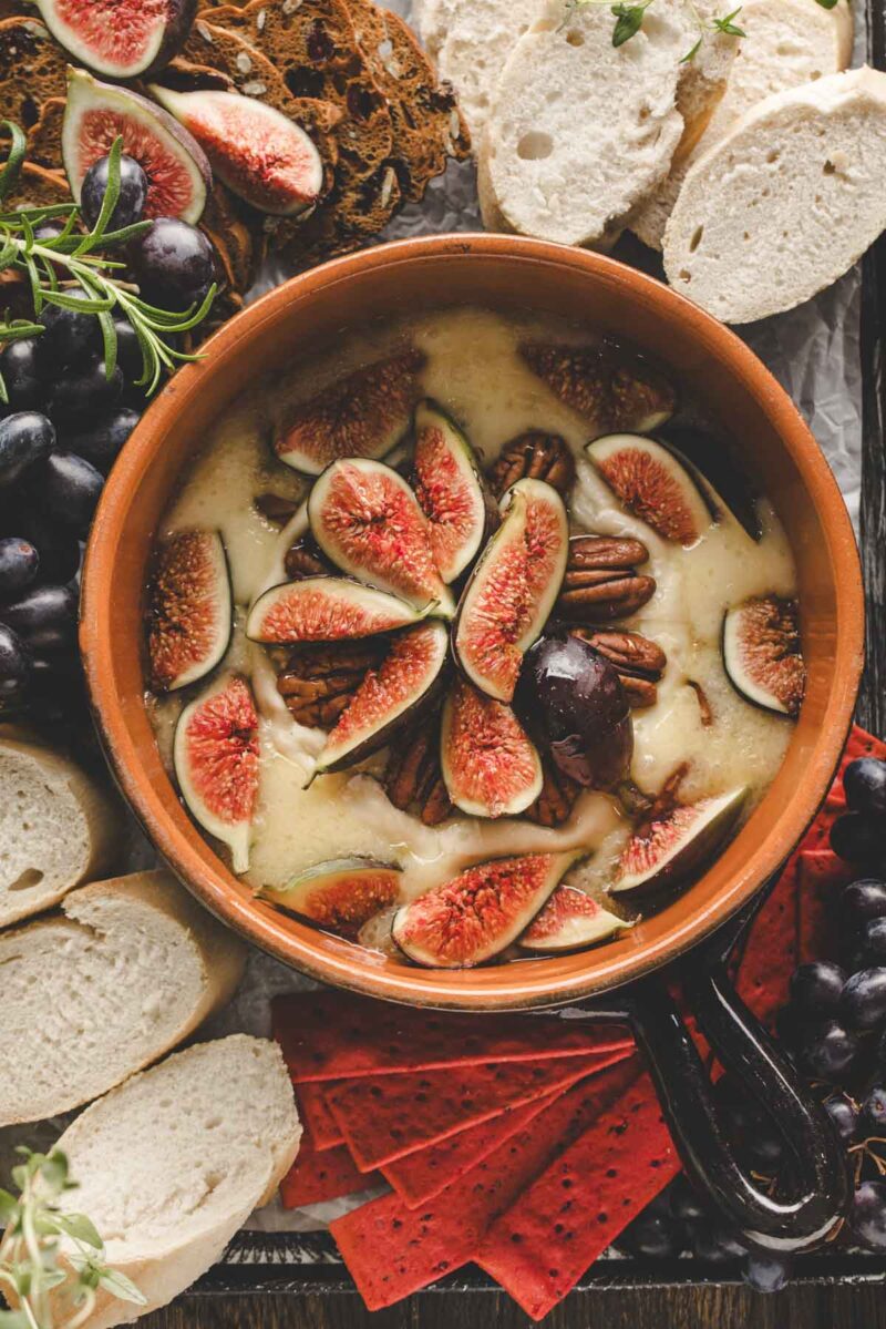 A dish of melted brie cheese topped with sliced figs and pecans, surrounded by sliced baguette, grapes, and crackers.
