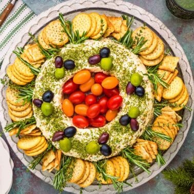 A round cheese spread topped with tomatoes and olives, garnished with herbs and surrounded by crackers, on a platter with pinecones and drinks nearby.