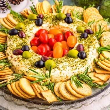 A round cheese and herb spread is garnished with tomatoes and olives, surrounded by crackers and rosemary sprigs on a glass plate.