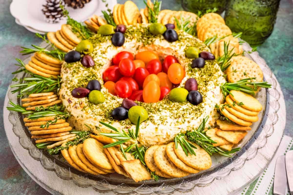 A round cheese and herb spread is garnished with tomatoes and olives, surrounded by crackers and rosemary sprigs on a glass plate.