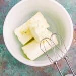 Three sticks of butter in a white mixing bowl next to an electric hand mixer on a green textured surface.