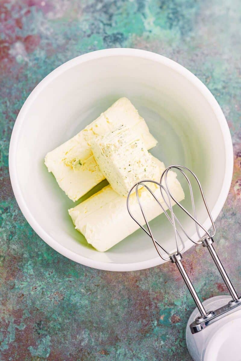 Three sticks of butter in a white mixing bowl next to an electric hand mixer on a green textured surface.