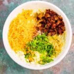 A white bowl contains shredded cheddar cheese, chopped green onions, crumbled bacon, and seasonings on a marbled green surface.