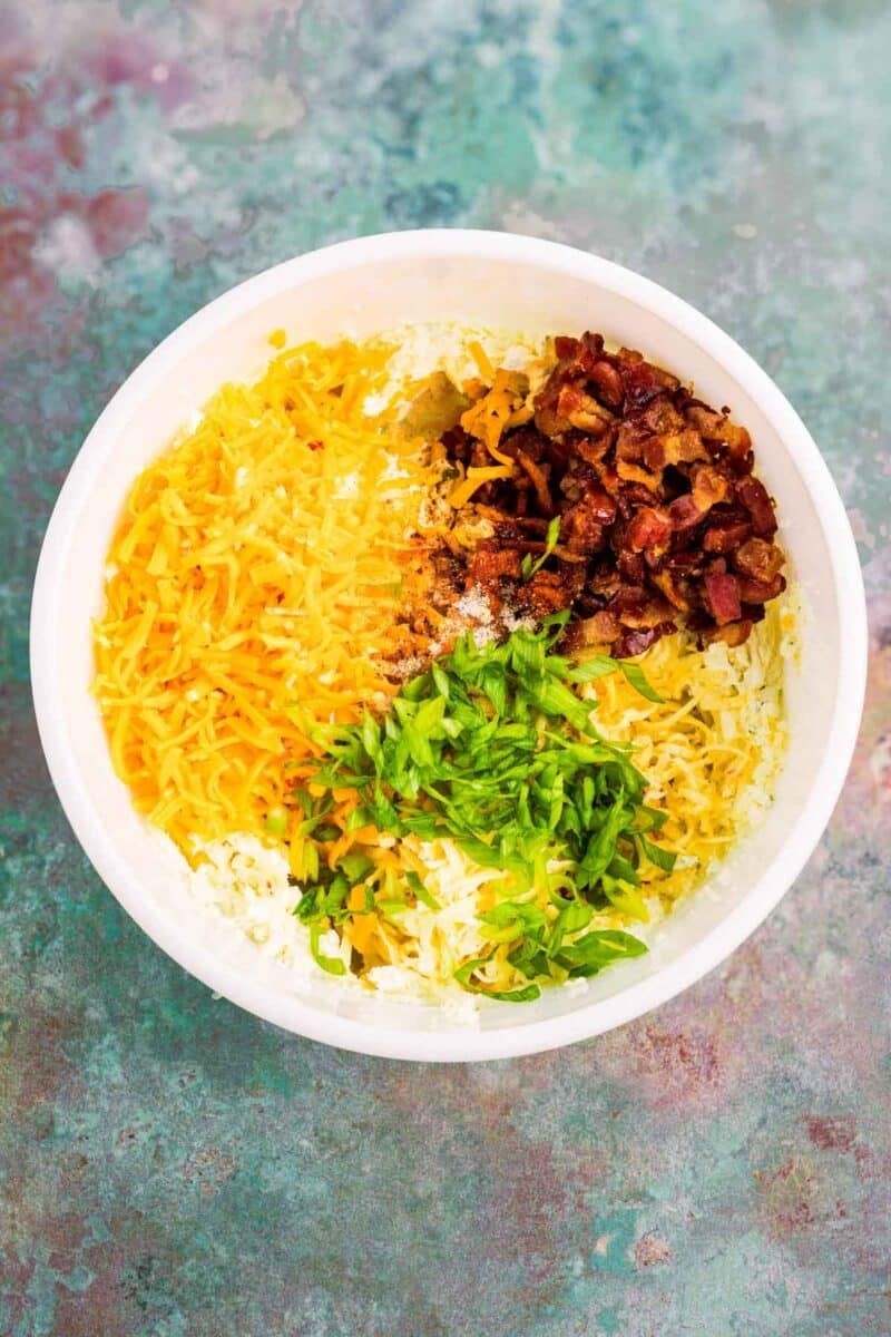A white bowl contains shredded cheddar cheese, chopped green onions, crumbled bacon, and seasonings on a marbled green surface.