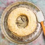 A ring-shaped cheese ball with visible herbs and spices sits on a clear glass platter next to a white spatula with a wooden handle.