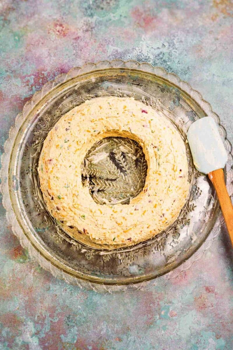 A ring-shaped cheese ball with visible herbs and spices sits on a clear glass platter next to a white spatula with a wooden handle.