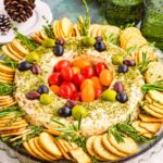 A festive cheese wreath topped with olives and cherry tomatoes, surrounded by crackers and garnished with herbs, sits on a glass platter.