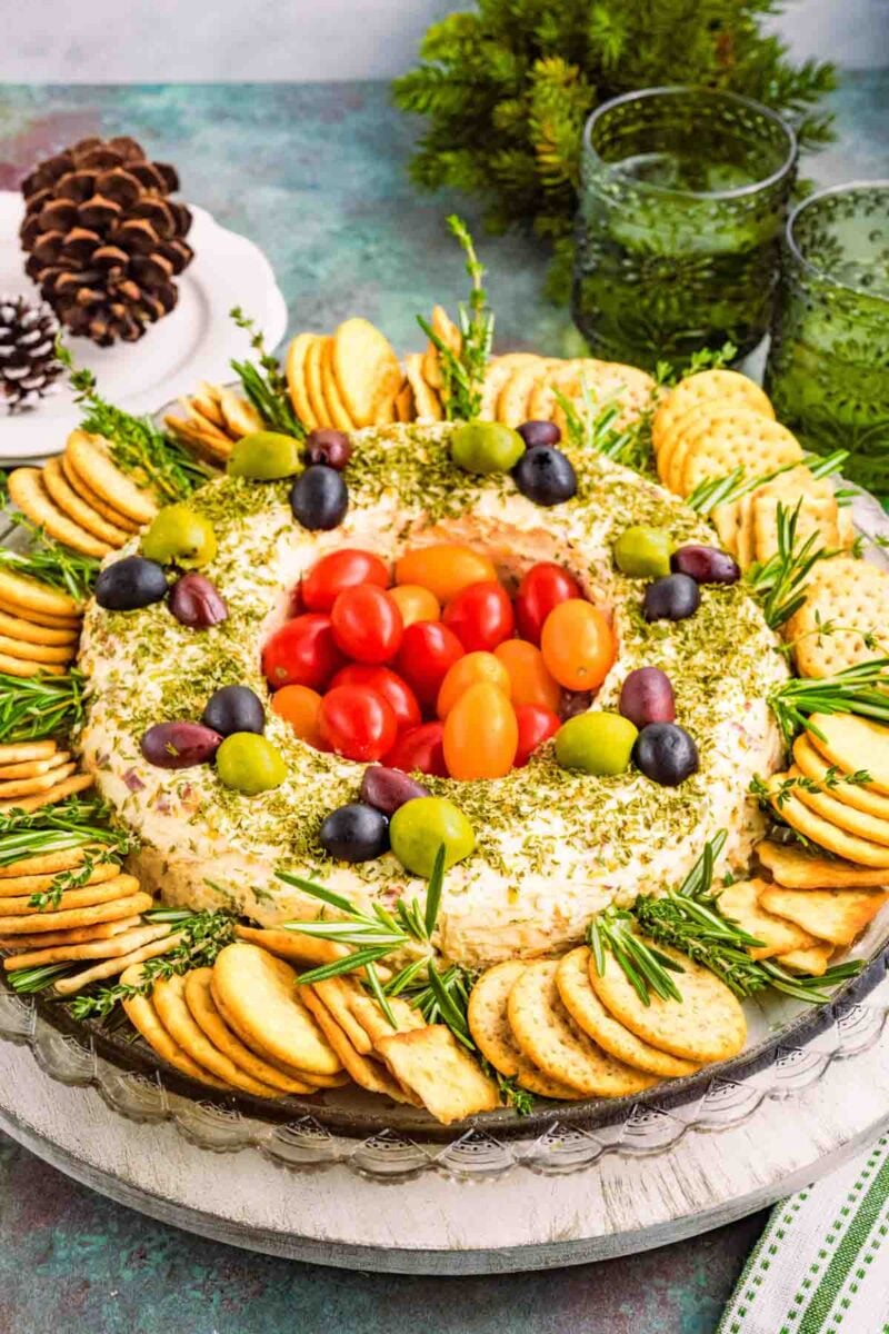 A festive cheese wreath topped with olives and cherry tomatoes, surrounded by crackers and garnished with herbs, sits on a glass platter.