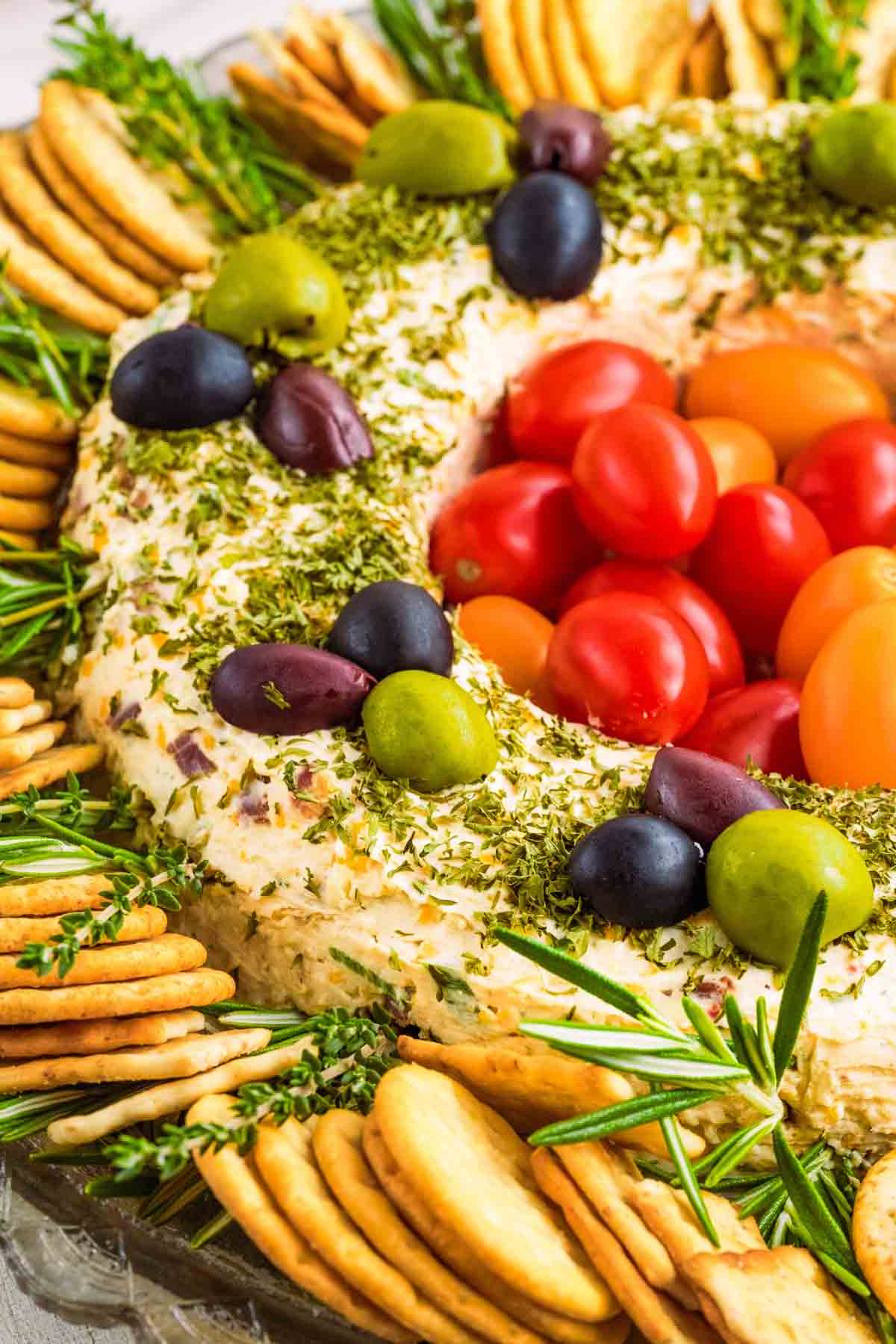 A festive cheese wreath topped with herbs, olives, and cherry tomatoes, surrounded by assorted crackers and sprigs of rosemary.