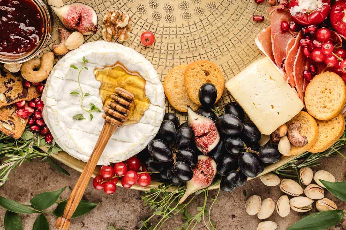 A platter with brie cheese topped with honey, black grapes, figs, crackers, prosciutto, red currants, pistachios, pomegranate seeds, cookies, and a jar of jam.