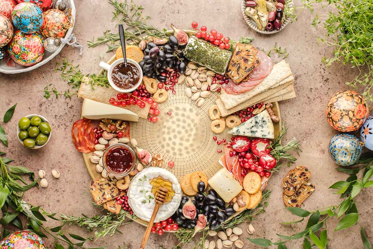 A circular cheese board with assorted cheeses, crackers, salami, grapes, nuts, honey, jams, and herbs, surrounded by painted eggs, olives, and greenery.