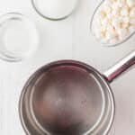 An empty saucepan, a glass bowl of sugar, a measuring cup of mini marshmallows, and a small bowl of water on a white surface.