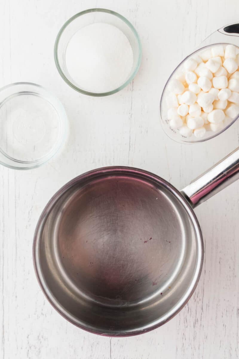 An empty saucepan, a glass bowl of sugar, a measuring cup of mini marshmallows, and a small bowl of water on a white surface.
