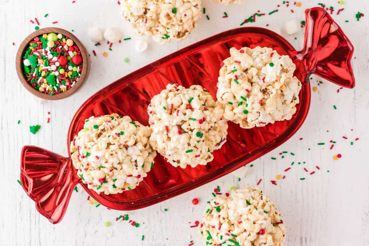 Four popcorn balls with colorful holiday sprinkles are arranged on a red candy-shaped tray, with a bowl of extra sprinkles nearby.