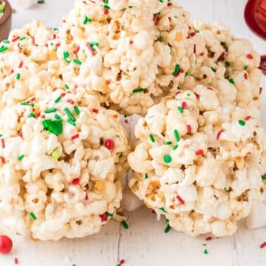Popcorn balls decorated with red, green, and white sprinkles are stacked on a white surface with marshmallows and more sprinkles scattered around.